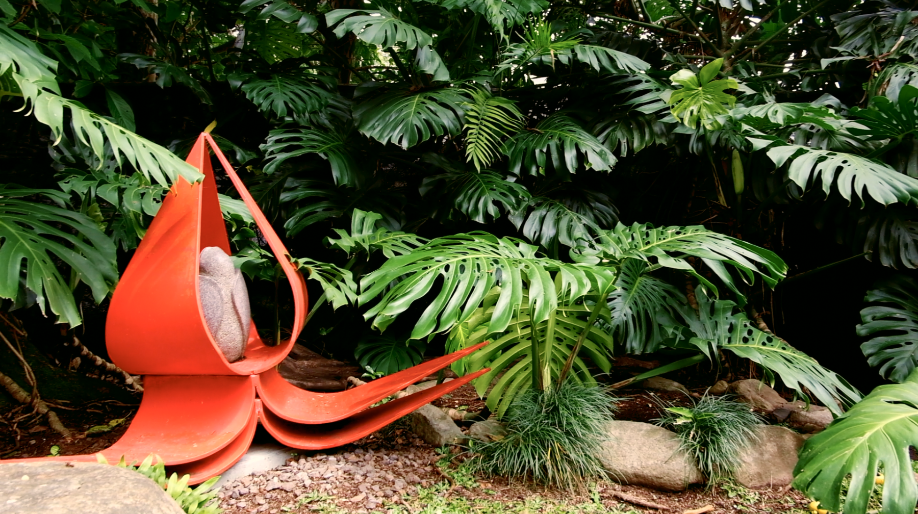 Una escultura de la flor de loto en rojo chroma que resalta por su tono saturado contra un fondo de naturaleza tropical verde. Escultor: José Sancho Benito